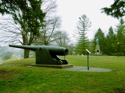 A historic cannon on a grassy area with trees, overlooking a statue in the background on a foggy day.