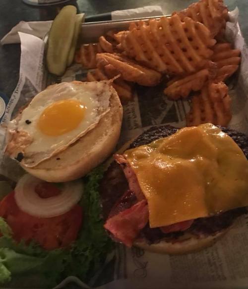 A burger with cheese, bacon, lettuce, tomato, and a fried egg, served with waffle fries and pickles on the side.