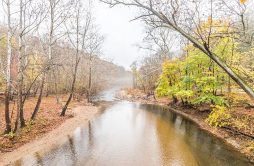A serene river flows through a foggy landscape, surrounded by autumn trees with yellow and orange leaves.
