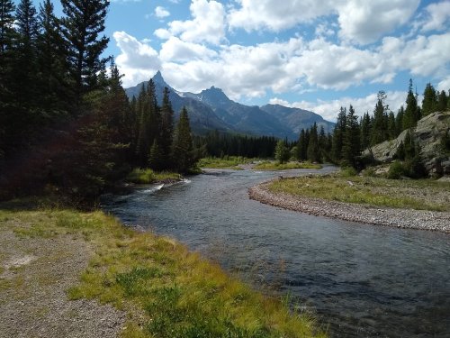 A winding river flows through a lush green landscape, surrounded by mountains and trees under a partly cloudy sky.