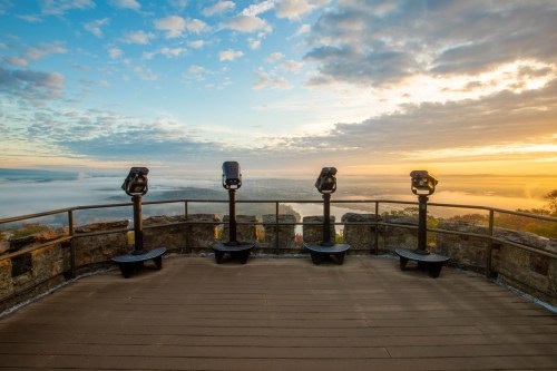 Viewpoint with binoculars overlooking a foggy landscape at sunrise, surrounded by clouds and a colorful sky.