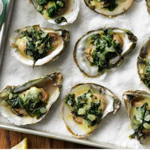 Baked oysters topped with greens and herbs on a bed of salt, arranged on a baking tray.