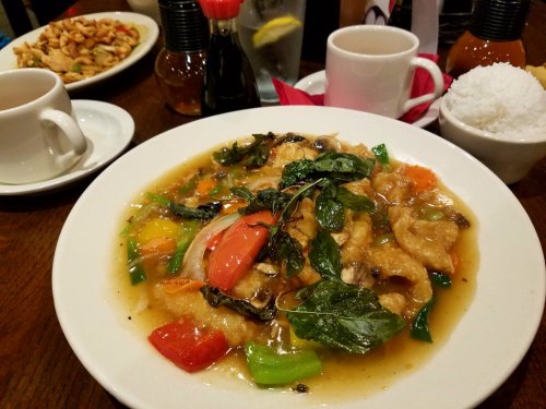 A plate of colorful stir-fried fish with vegetables and herbs, served with rice and drinks in a restaurant setting.