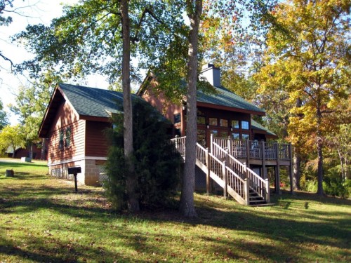 A cozy wooden cabin surrounded by trees, featuring a deck and stairs leading to the entrance.