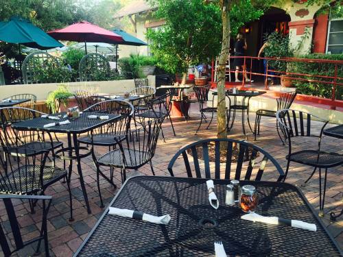 Outdoor dining area with black metal tables and chairs, surrounded by greenery and colorful umbrellas.
