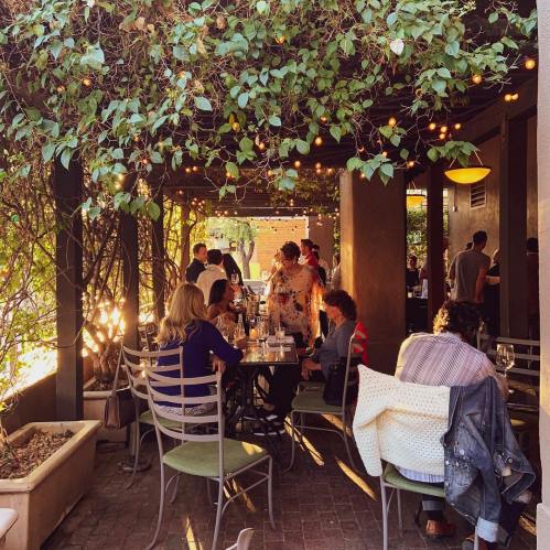 A lively outdoor dining area with people enjoying meals under a canopy of greenery and string lights.