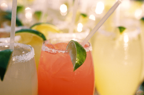 Colorful cocktails with lime and lemon garnishes, served in glasses with straws and salt rims.