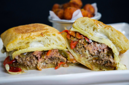 A close-up of a sliced sandwich filled with meat, peppers, and cheese, served with a side of tater tots.