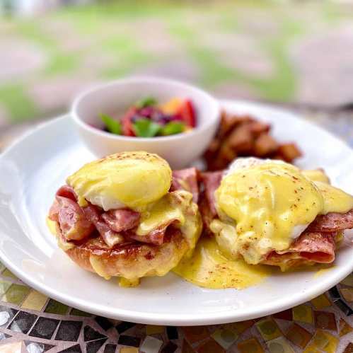 Two eggs Benedict on English muffins with ham and hollandaise sauce, served with a side of fruit and potatoes.