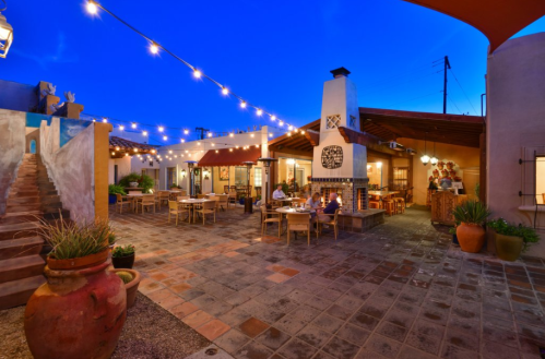 A cozy outdoor dining area with string lights, tables, and a fireplace, set against a twilight sky.