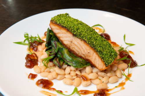 A beautifully plated salmon fillet topped with green herb crust, served on a bed of greens and white beans with sauce.