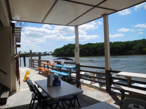 View from a covered patio overlooking a river, with tables and chairs, and lush greenery in the background.