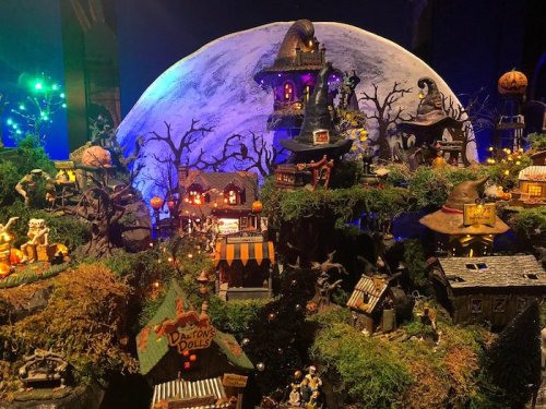 A whimsical Halloween village scene with miniature houses, trees, and a large moon backdrop, illuminated with colorful lights.