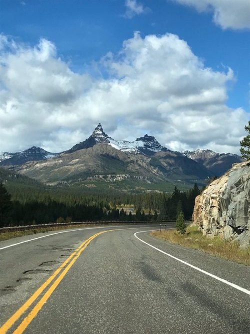 A winding road leads through a mountainous landscape with snow-capped peaks and a blue sky dotted with clouds.