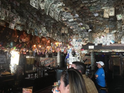 A lively bar interior with walls and ceiling covered in dollar bills and various decorations, patrons enjoying drinks.