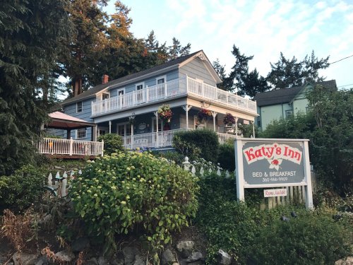 A charming gray bed and breakfast with a white porch, surrounded by greenery and flowers, featuring a sign for Katy's Inn.