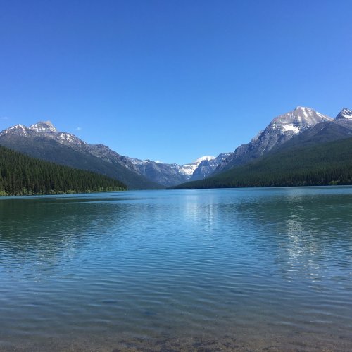 A serene lake surrounded by mountains under a clear blue sky, with lush green trees lining the shore.
