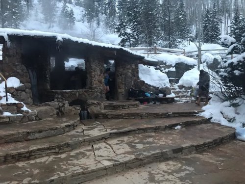 A rustic stone cabin surrounded by snow-covered trees, with steam rising and people nearby.