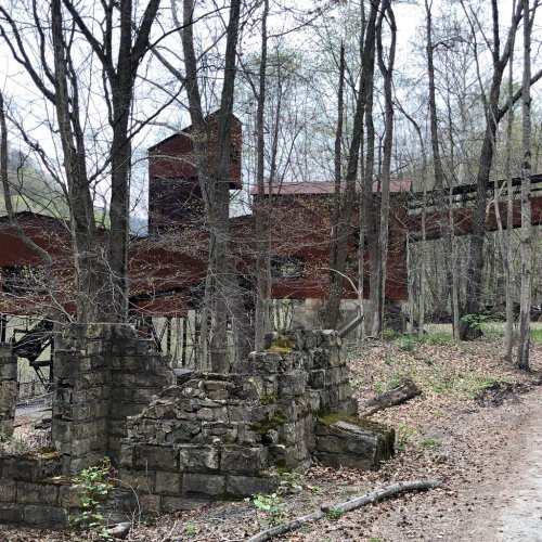 Abandoned red structure among trees, with stone ruins in the foreground and a forested landscape in the background.