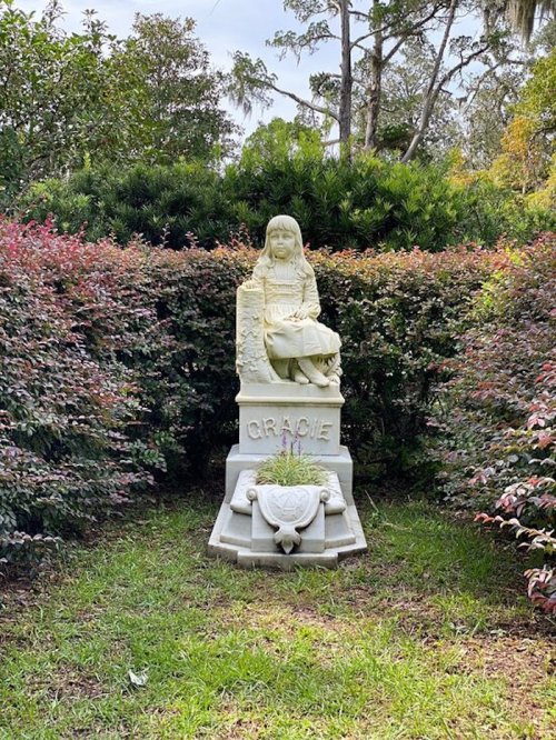 A stone statue of a girl named Gracie sits surrounded by lush greenery and colorful bushes.