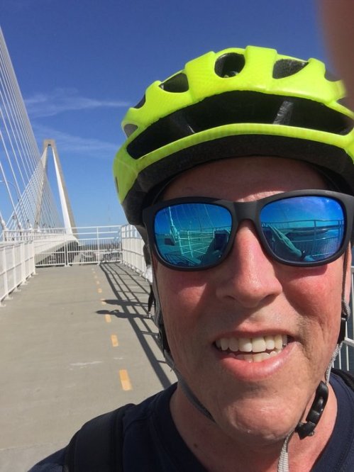 A person wearing a bright yellow helmet and sunglasses smiles for a selfie on a bridge with blue skies in the background.