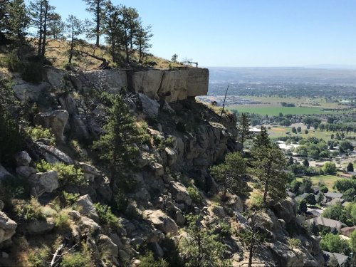 A rocky cliff with trees overlooks a valley and distant hills under a clear blue sky.