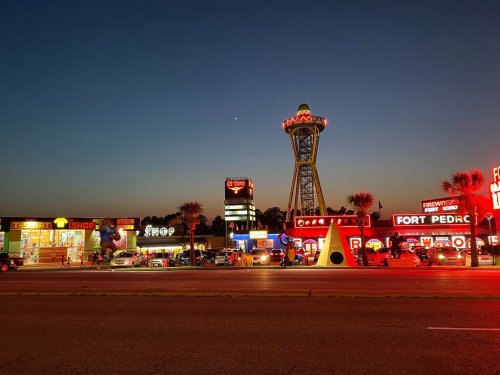 Colorful shops and a tall tower illuminated at dusk, with cars parked along a busy street.