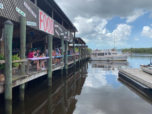 A waterfront restaurant with diners on a deck, boats docked nearby, and a cloudy sky above.