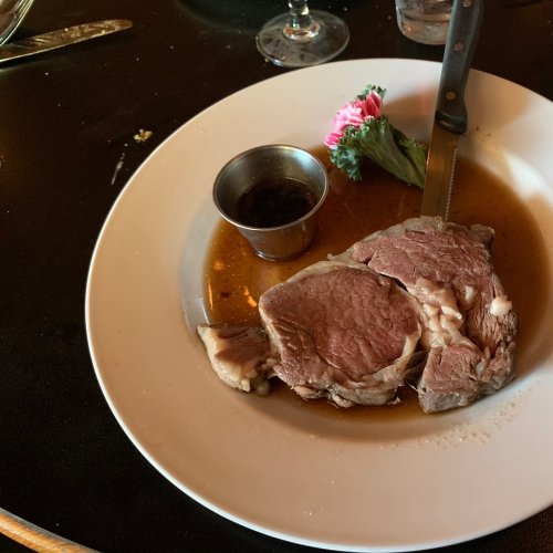 A plate with a slice of prime rib, a small cup of sauce, and a pink flower garnish on a black table.