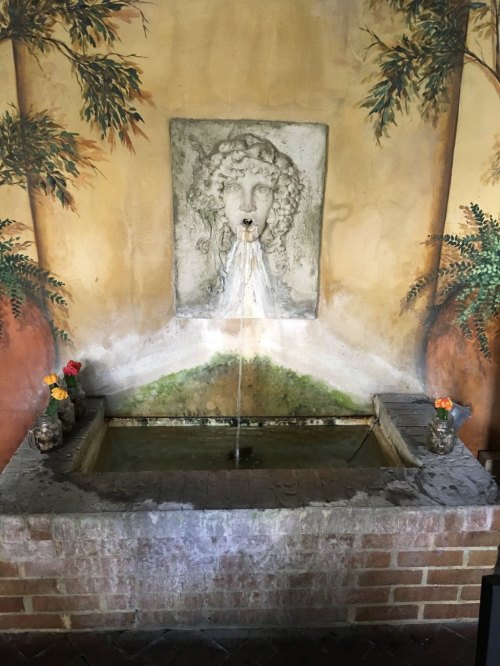 A stone fountain with a lion's head spout, surrounded by a mural of trees and flowers, set against a brick wall.