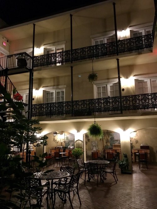 A dimly lit courtyard with tables and chairs, surrounded by balconies and greenery, creating a cozy evening atmosphere.