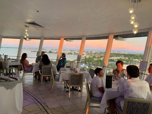 A restaurant interior with diners enjoying a sunset view over water and cityscape through large windows.
