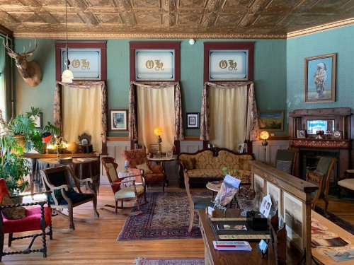 A vintage parlor with ornate decor, plush seating, plants, and warm lighting, featuring a mix of antique furniture.
