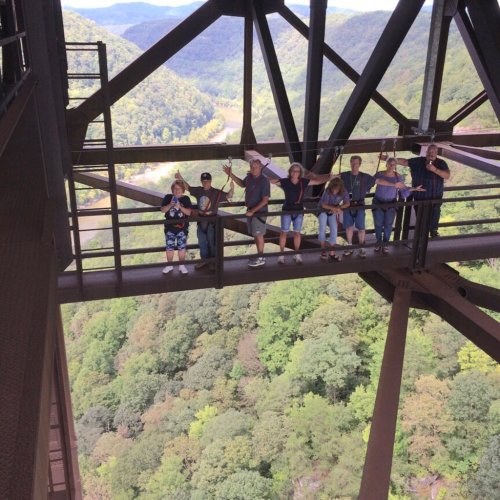 A group of people stands on a metal structure overlooking a lush green valley and mountains in the background.