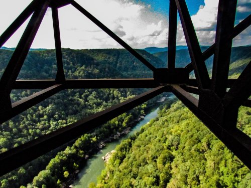 View of a lush green valley and river framed by the structure of a bridge, with mountains in the background and a blue sky.