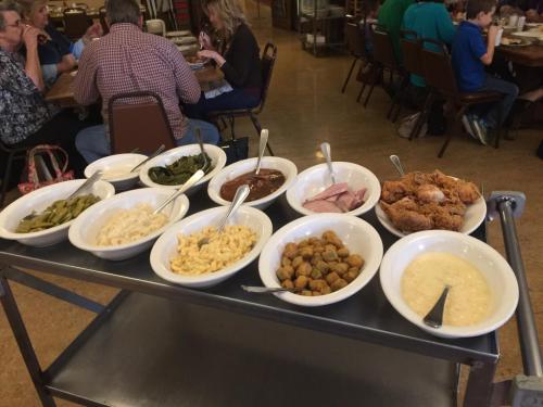 A serving cart with various dishes: greens, macaroni, ham, fried chicken, and more, in a busy dining setting.