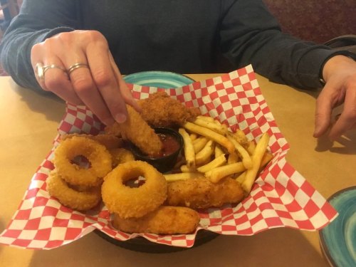 A hand dipping an onion ring into a small bowl of sauce, surrounded by fries and other fried foods on a checkered plate.
