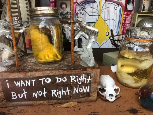 A wooden sign reads "I WANT TO DO RIGHT But NOT RIGHT NOW," beside jars with yellow contents and a small skull.
