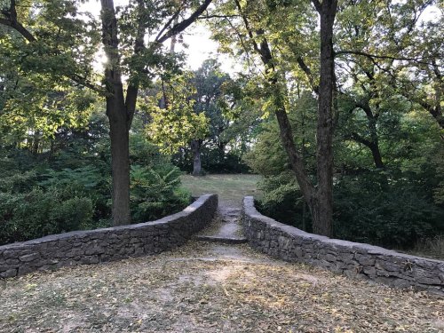 A stone bridge leads through a wooded area, surrounded by trees and dappled sunlight.