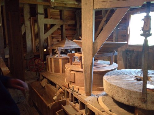 Interior of a historic mill featuring wooden machinery and large stone grinding wheels. Natural light from a window.