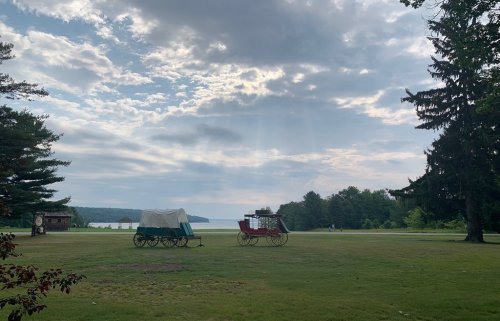 Two covered wagons on a grassy field with a lake in the background and cloudy skies above.