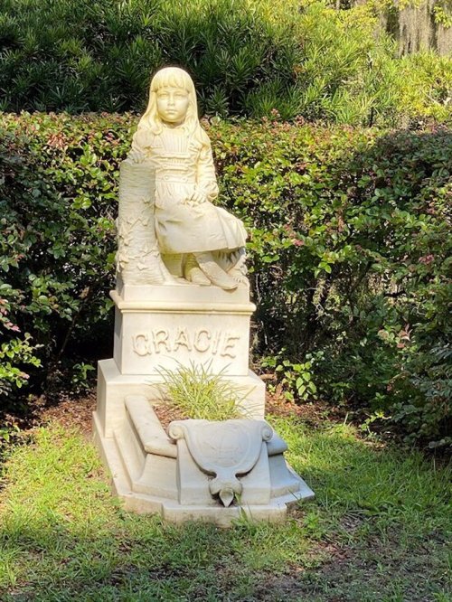 A stone statue of a girl named Gracie sitting on a pedestal, surrounded by greenery in a garden setting.