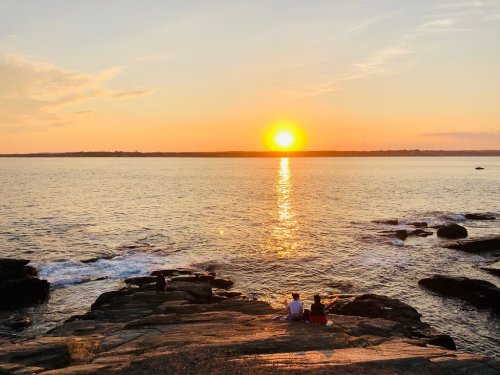 A couple sits on rocky shore watching a vibrant sunset over the calm ocean, with golden reflections on the water.