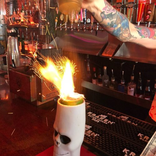 A bartender pours a flaming drink into a skull-shaped glass, creating sparks and flames in a vibrant bar setting.