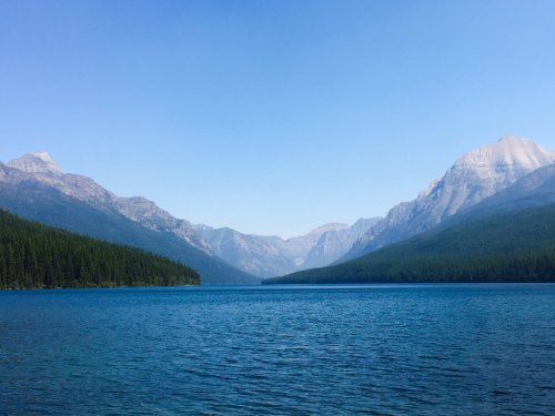 A serene lake surrounded by mountains and dense forest under a clear blue sky.