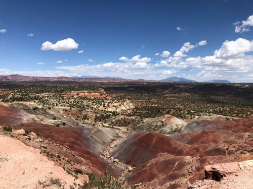 Vast desert landscape with colorful rock formations, rolling hills, and a blue sky dotted with fluffy white clouds.