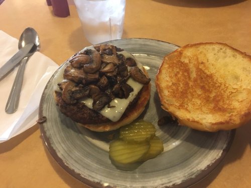 A burger topped with melted cheese and sautéed mushrooms, served with pickles and a toasted bun on a plate.