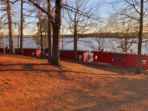Red train cars lined up near a lake, surrounded by bare trees and a warm, golden sunset.