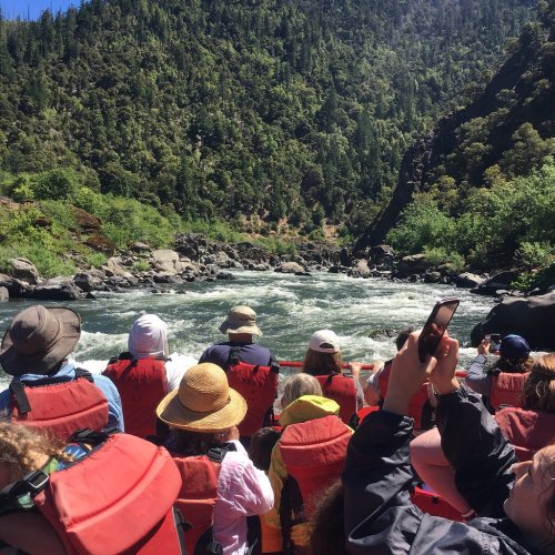 A group of people in life jackets on a raft, navigating a river surrounded by lush greenery and rocky terrain.