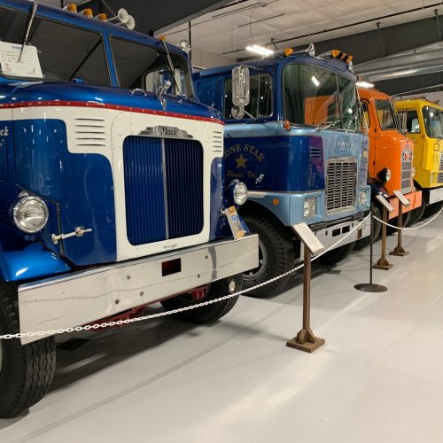 A row of vintage trucks in a museum, showcasing various colors and styles, with informational plaques beside each.